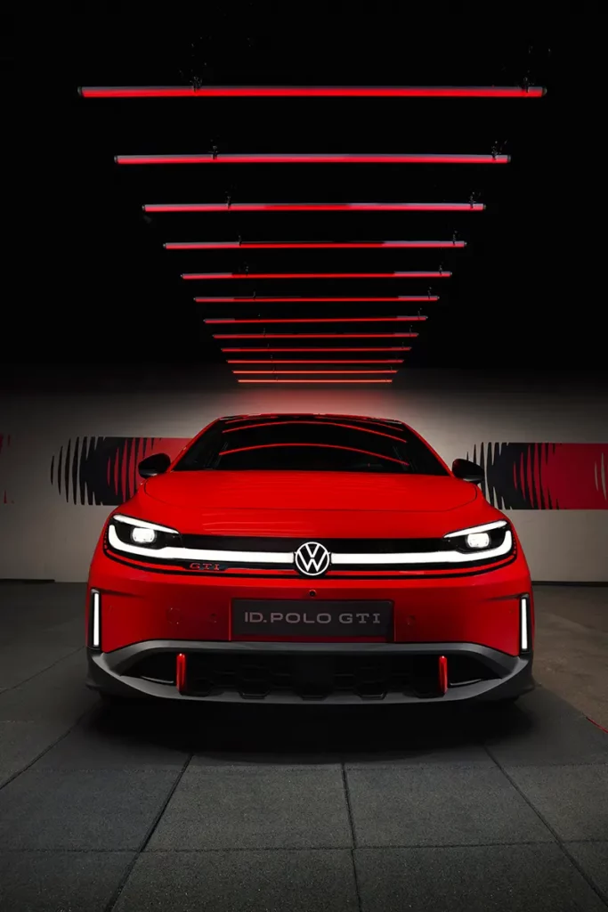 Vue de face au grand-angle d'une Volkswagen ID. Polo GTI électrique rouge, centrée sur des lignes de symétrie au sol et des néons rouges au plafond avec logos et phares allumés.