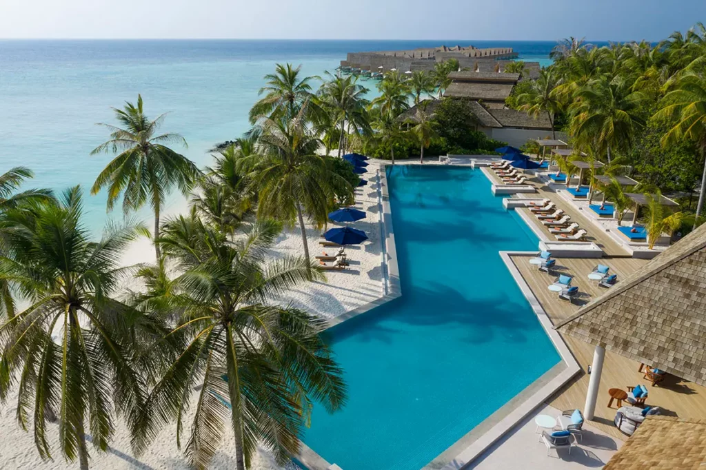 Vue aérienne en plongée d'Emerald Faarufushi montrant la piscine principale, les toits des Beach Villas et les villas sur pilotis s'étendant dans l'Océan Indien.