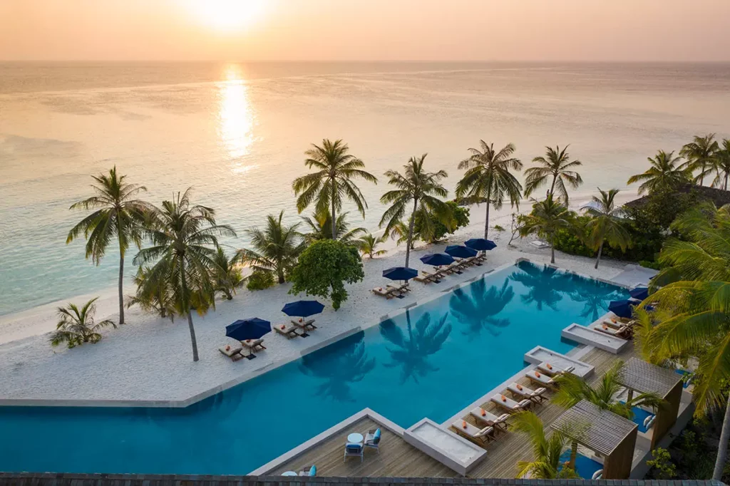 Vue aérienne de la grande piscine et des infrastructures du resort Emerald Faarufushi aux Maldives lors du coucher de soleil sur l'Océan Indien.