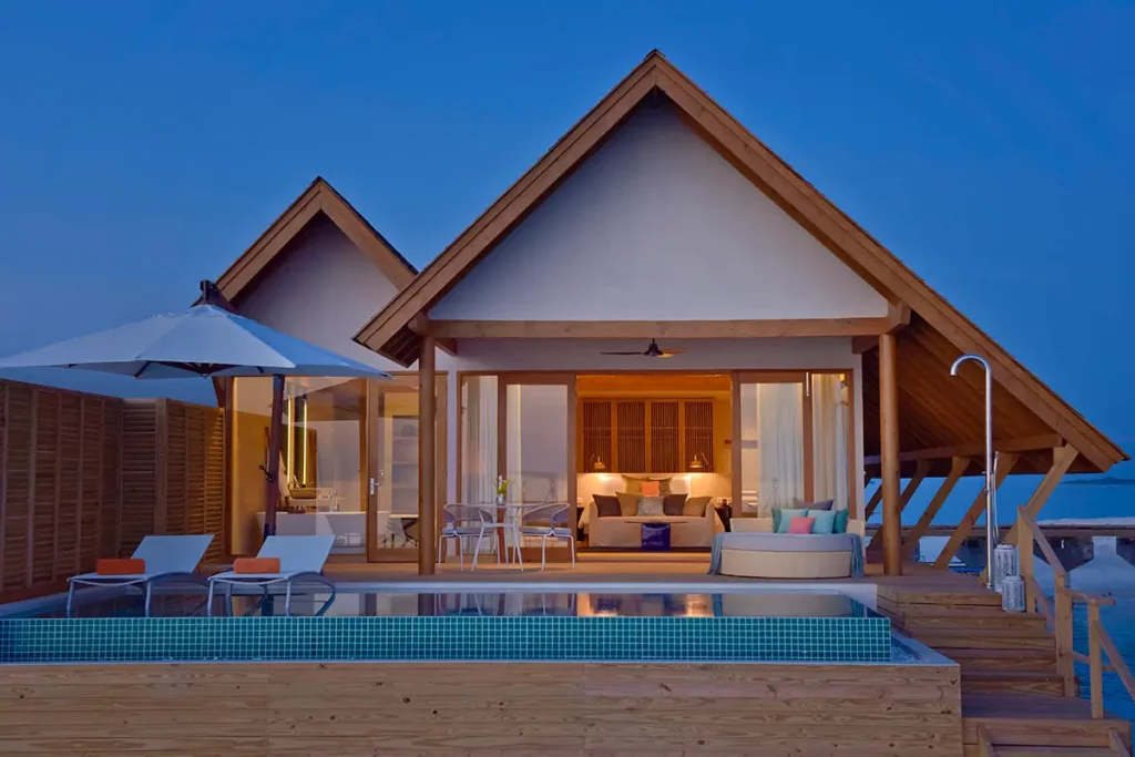Vue depuis l'océan d'une Water Villa à Emerald Faarufushi à l'heure bleue, montrant la piscine privée, l'escalier d'accès et l'intérieur de la suite illuminé.
