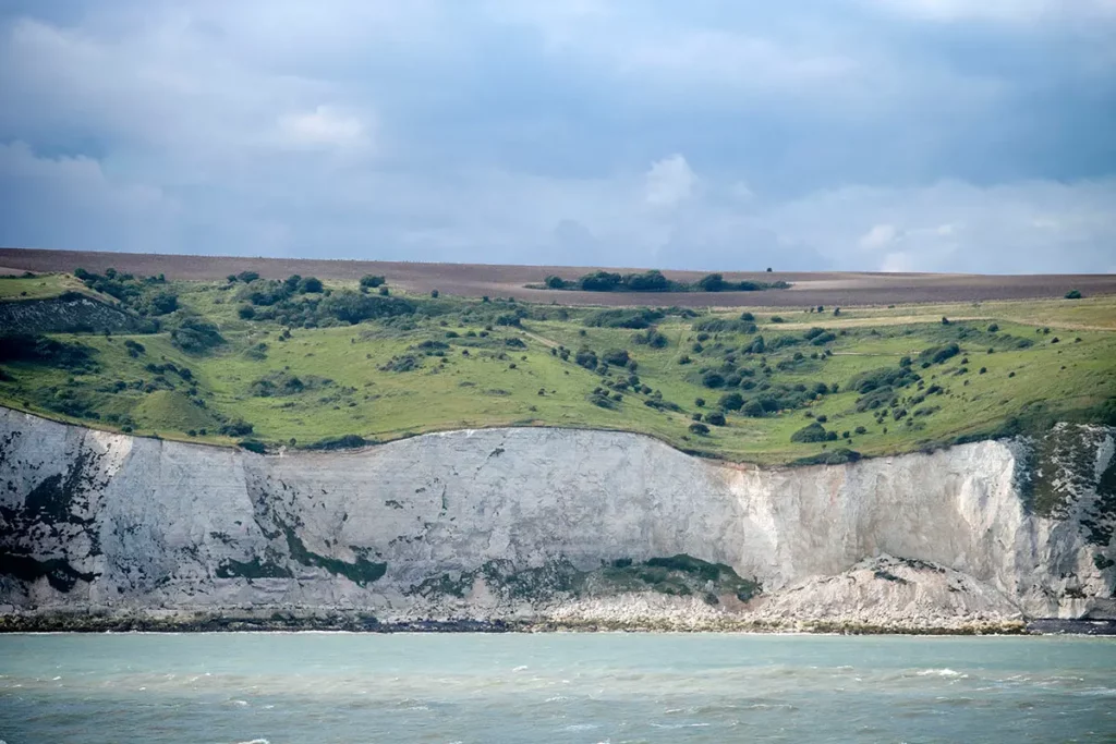 Les falaises de Douvres vues du bateau, montrant les strates de la roche blanche, de la végétation verte et d'un champ de terre brune labouré sous le ciel.