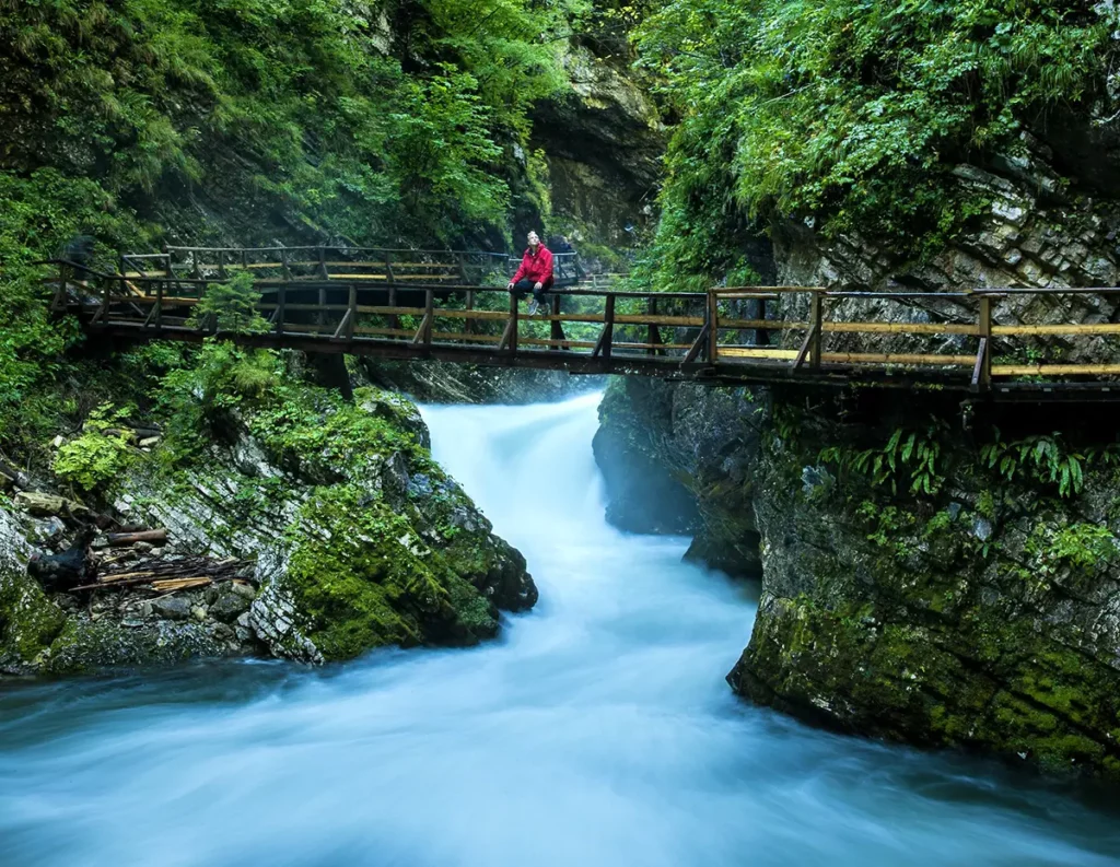 Un homme minuscule debout sur une longue passerelle en bois qui serpente au-dessus d'une rivière floutée par une pose longue dans les gorges étroites de Mostnica, illustrant la grandeur de la nature sauvage lors d'un road-trip en Slovénie.
