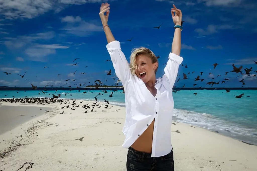 Julie Taton souriante en chemisier blanc et montre Baume et Mercier sur un banc de sable aux Maldives proche du Kanuhura, entourée d'oiseaux marins au Kanuhura.