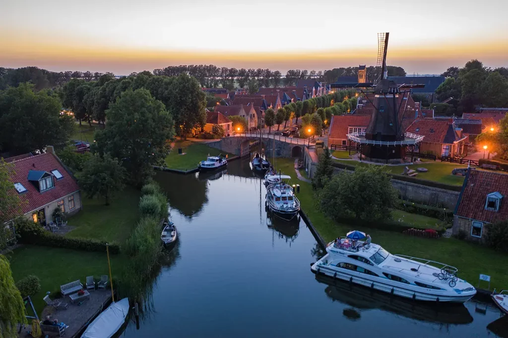 Vue aérienne par drone en début de nuit du modèle Le Boat Elegance amarré le long d'un canal à Sloten, dans le Friesland aux Pays-Bas, avec le moulin historique De Kaai éclairé au crépuscule.