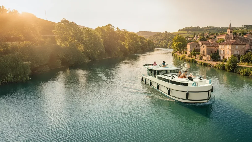 Vue aérienne au grand angle d'un bateau Le Boat naviguant à contre-jour sur un canal en France dans une lumière dorée de fin de journée, avec un village historique en vieilles briques sur la berge droite.