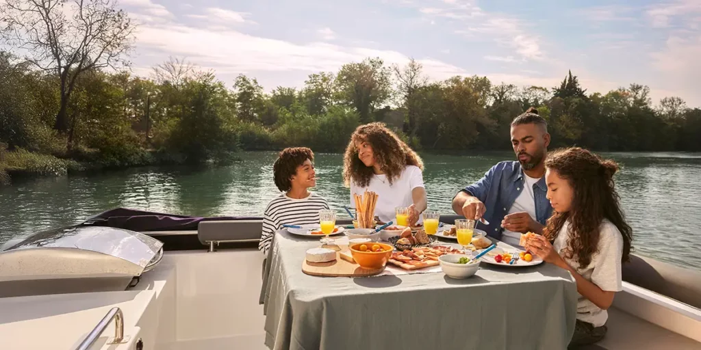 Une famille avec deux enfants partage un repas léger autour de la table sur le pont arrière d'un bateau Le Boat, amarré le long d'une belle voie navigable.