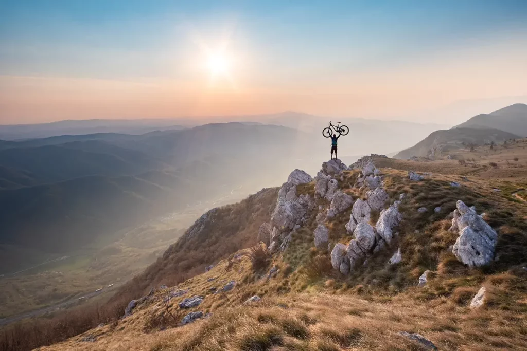 Un homme minuscule au sommet du plateau de Nanos lève son VTT en victoire au-dessus de la vallée de Vipava au coucher du soleil, lors d'un road-trip en Slovénie.