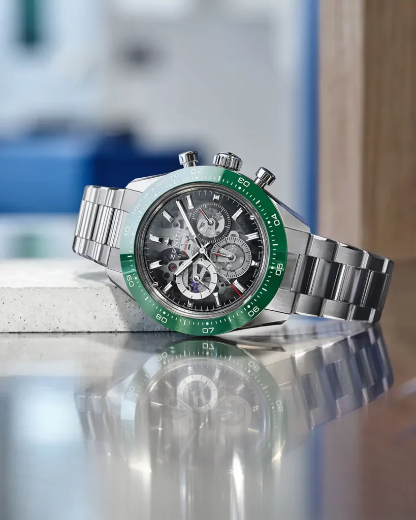 Montre Zenith Chronomaster Sport Skeleton acier à lunette verte couchée sur une plaque réfléchissante et un socle en pierre blanche.