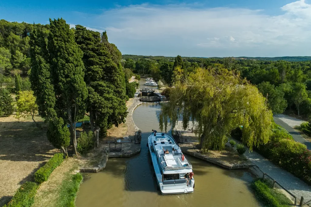 Bateau Le Boat Horizon 5 manœuvrant à l'approche d'une écluse sur le canal du Midi, entouré d'une végétation dense, avec d'autres bateaux en attente en arrière-plan.