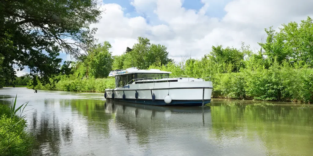 Bateau Le Boat Horizon 5 naviguant paisiblement sur un canal bordé d'une végétation dense, vu depuis la berge, illustrant l'expérience du slow travel.