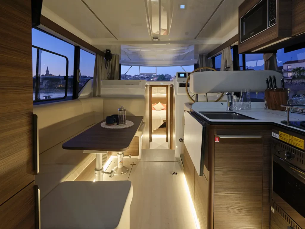 Ambiance chaleureuse dans le salon et la cuisine d'un bateau Liberty Le Boat en début de soirée, avec vue sur les lumières d'un village au loin.