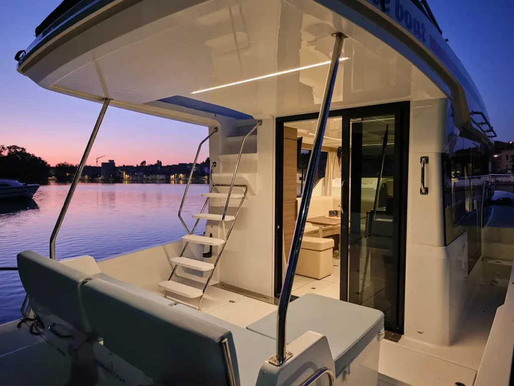 Vue arrière du bateau Liberty Le Boat en début de nuit, mettant en valeur les coussins confortables, l'accès au deck supérieur et l'ambiance lumineuse chaleureuse du bateau.