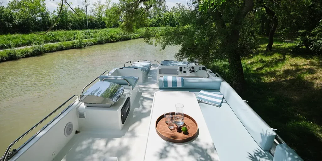 Pont supérieur aménagé d'un bateau Liberty Le Boat avec table prête à être dressée, poste de pilotage extérieur et barbecue, amarré sur un canal verdoyant.