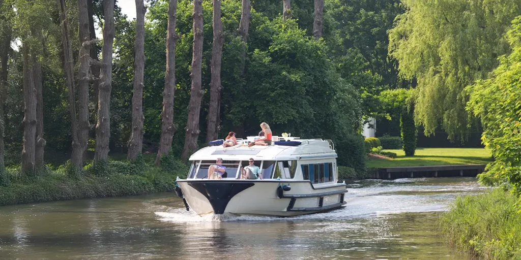 Bateau Le Boat naviguant face à l'objectif sur un canal en Belgique, bordé par la végétation et les arbres typiques des paysages de Flandre.