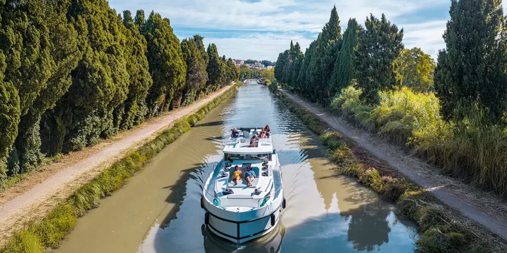 Bateau Le Boat naviguant sur le canal du Midi près de Carcassonne, avec un couple se relaxant sur le pont avant et le pilote à la barre extérieure, dans un paysage aux berges naturelles.