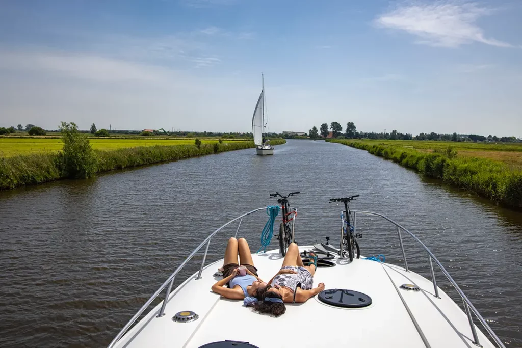 Détente sur le sun deck d'un bateau Le Boat naviguant sur un canal en Hollande, au milieu des champs, croisant un voilier en arrière-plan.