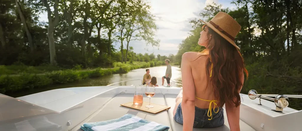 Femme en bikini se relaxant sur le pont supérieur d'un bateau Le Boat Horizon 5 sur le canal du Midi, avec un verre de vin rosé, tandis que des passagers discutent à la proue.