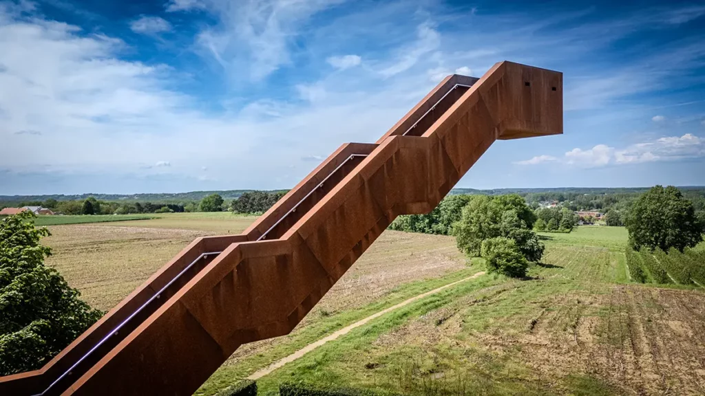 Vlooybergtoren en cadrage horizontal formant une diagonale en acier Corten traversant l'image entre campagne du Hageland et ciel bleu