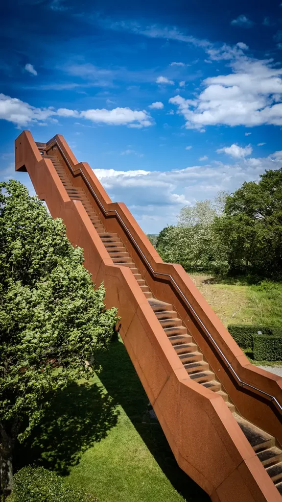 Vue latérale de la Vlooybergtoren par drone à mi-hauteur avec sa structure en acier Corten dominant un arbre voisin et les champs du Hageland