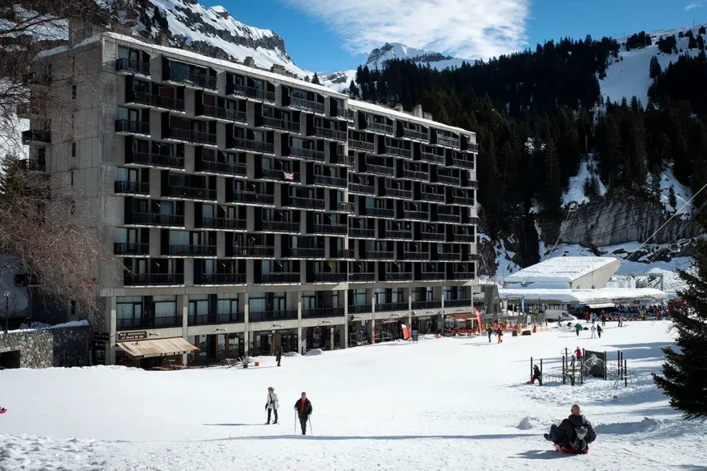 Photo horizontale montrant le dernier bâtiment de style Bauhaus sur le front de neige de Flaine avec des promeneurs et une personne en luge hors période de vacances