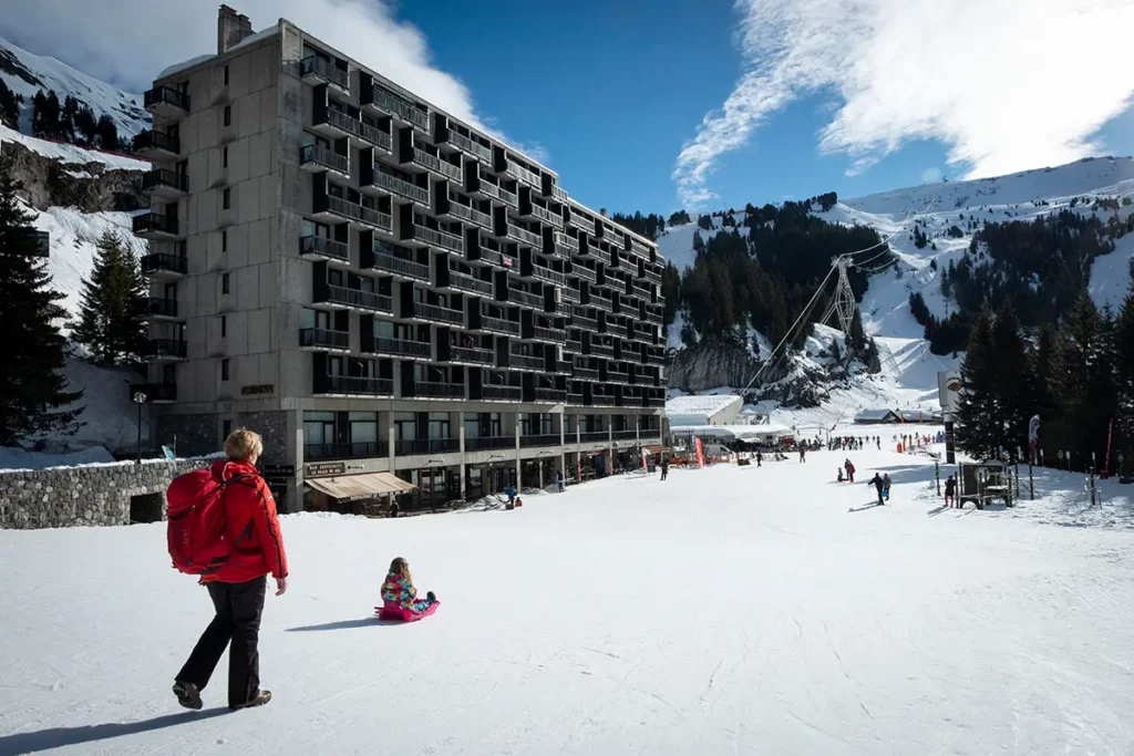 une femme marchant sur le front de neige de Flaine en suivant sa petite fille sur une luge avec les bâtiments Bauhaus et le décor minéral en arrière-plan
