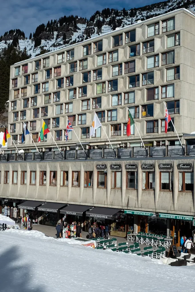 gros plan vertical sur un immeuble Bauhaus depuis le front de neige de Flaine avec ses commerces, ses fenêtres en bois brun et son balcon orné de drapeaux