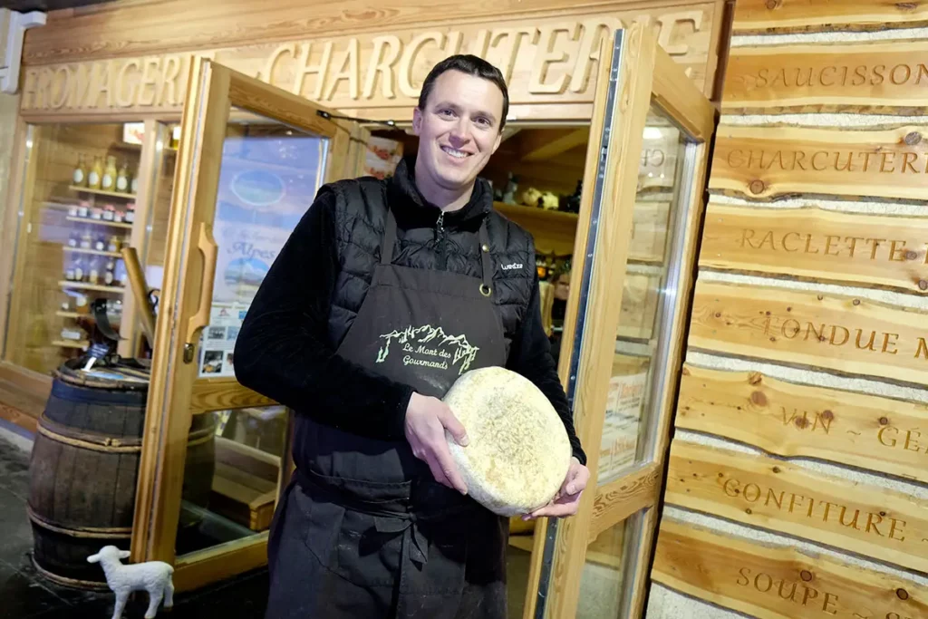 Éric Covemacker présentant une meule de fromage devant sa boutique Le Mont des Gourmands à Orcières Merlette 1850 dans les Hautes-Alpes