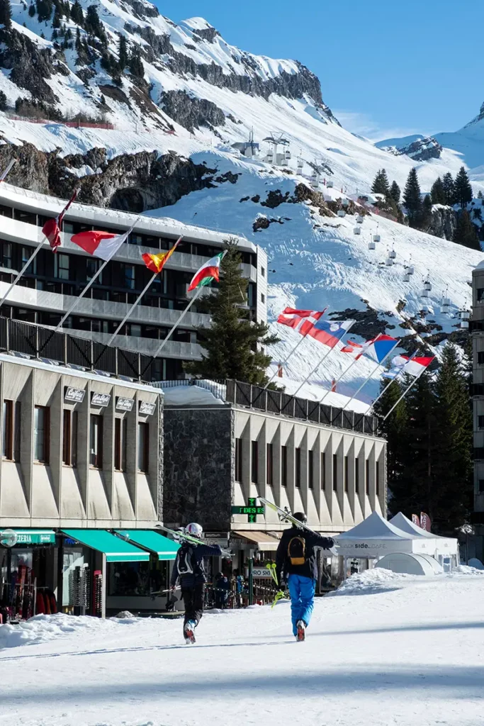 skieurs de dos marchant sur le front de neige avec leurs skis sur l'épaule vers les remontées mécaniques de Flaine sous un soleil radieux
