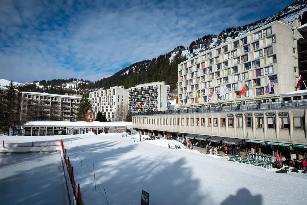 vue panoramique horizontale du front de neige de Flaine avec la patinoire au premier plan, les bâtiments Bauhaus de Marcel Breuer, les sapins enneigés et la sculpture colorée de Vasarely